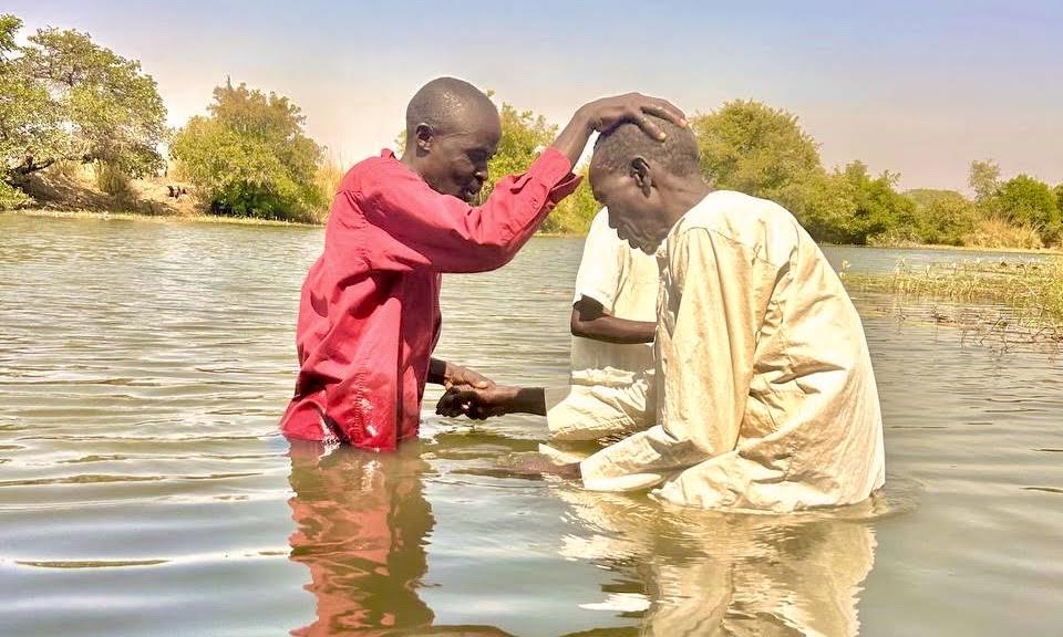 A person with hand placed on another person's head in pray. Both standing in water