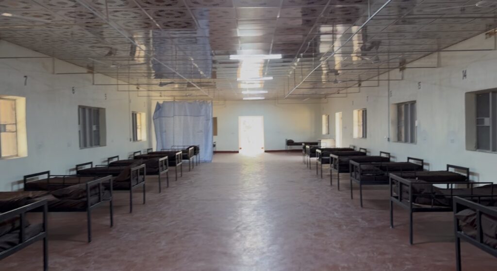 A large, empty hospital ward with rows of metal-framed beds aligned on both sides of a central aisle. The room features a reflective ceiling, multiple windows, and a bright doorway at the far end, suggesting a clean and orderly environment.