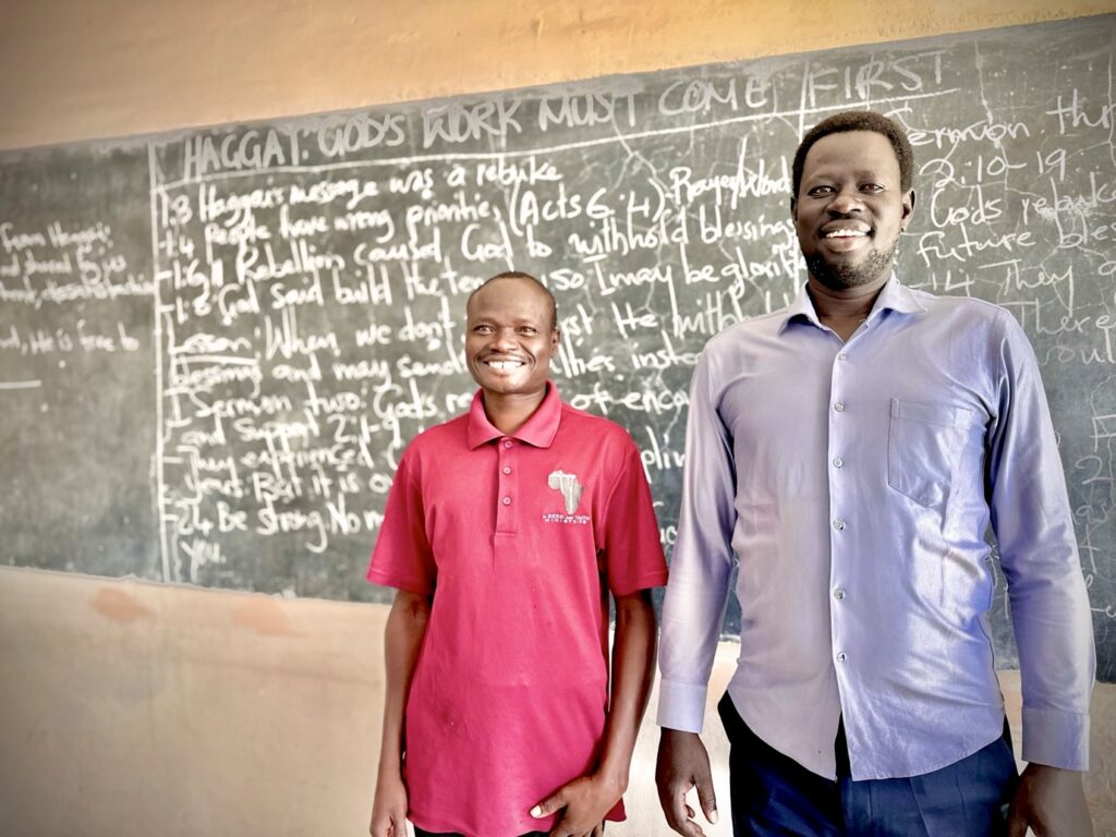 Two men standing in front of a classroom chalkboard covered with handwritten notes about a biblical topic.